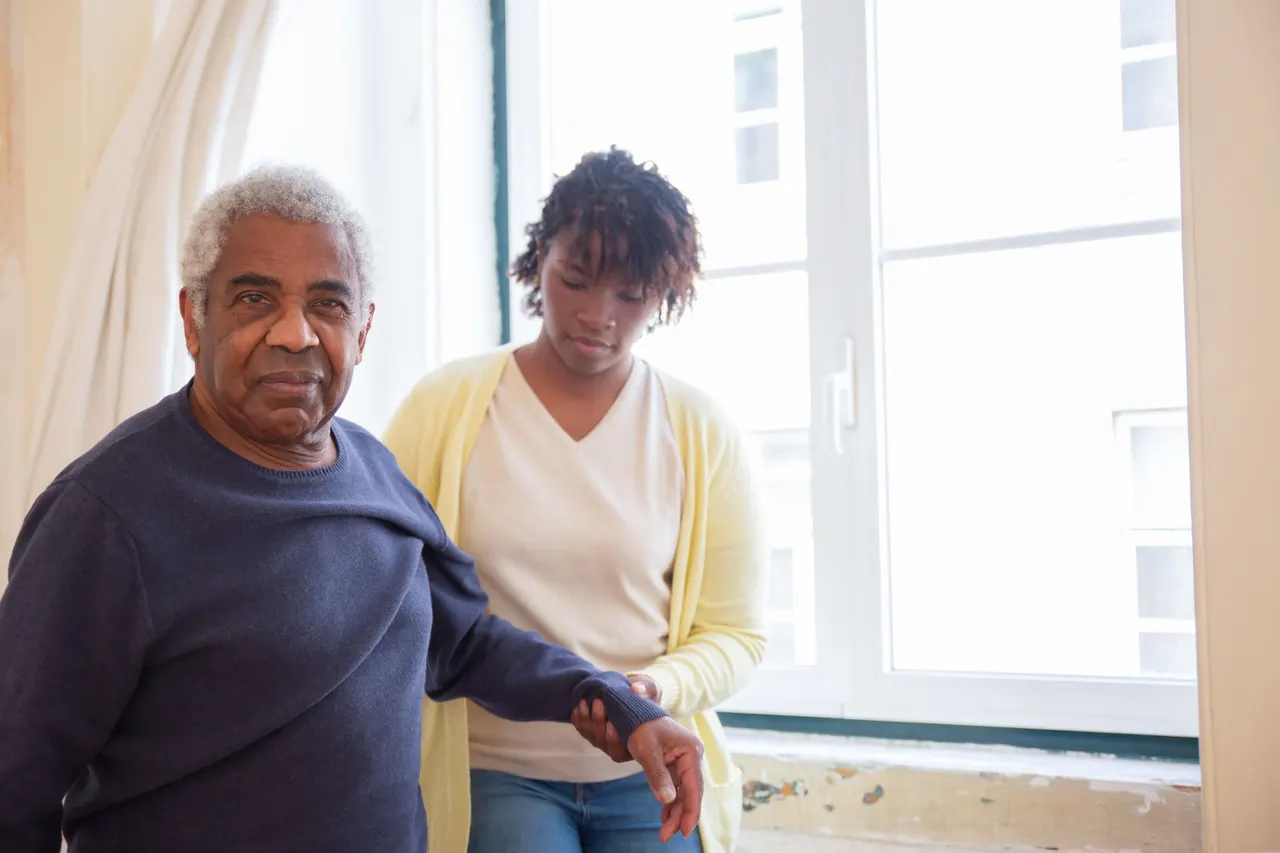 A caregiver supporting an older man's arm as he stands by a bright window