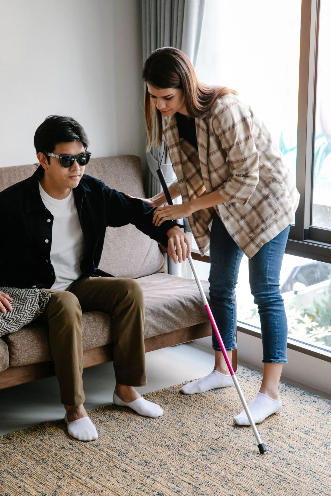 A support worker gently helping a man with a white cane rise from the sofa at home