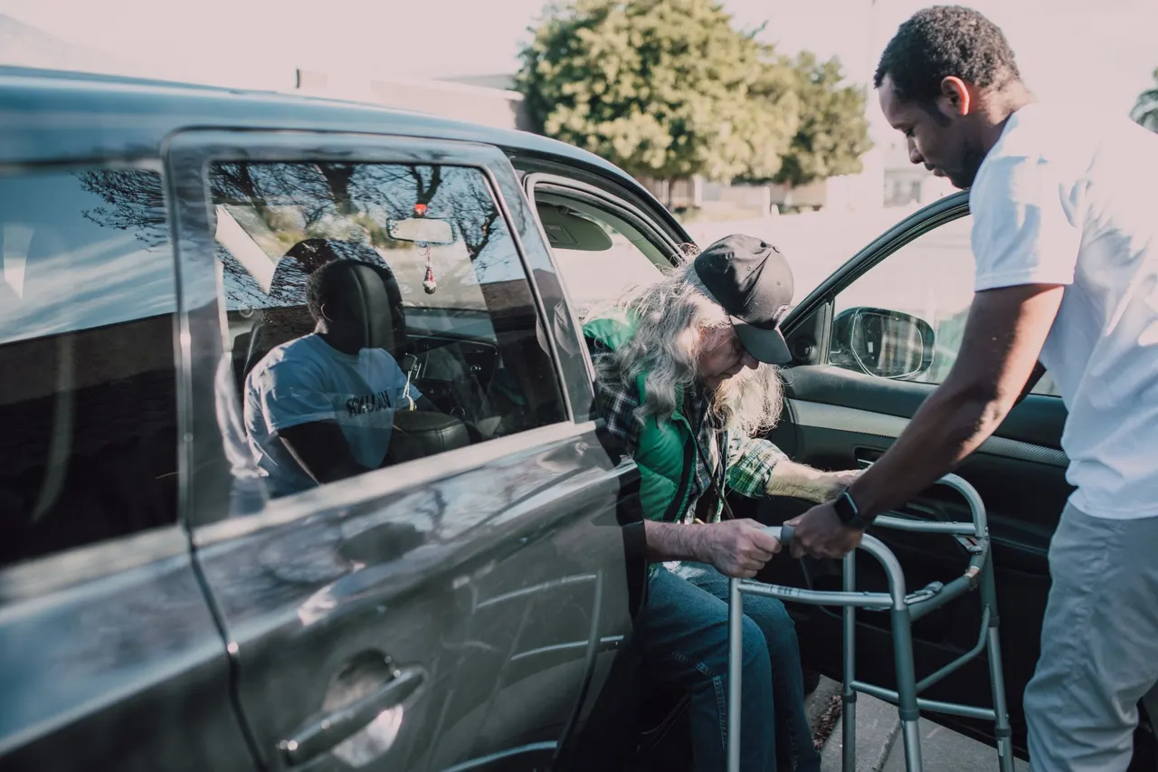 A support worker steadying a walker as an older man steps out of a car