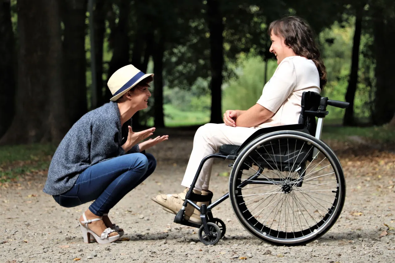 Two friends laughing together in a park, one using a wheelchair